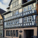 Fachwerkhaus im Landkreis Hersfeld-Rotenburg. Das mehrstöckige Gebäude besitzt eine aufwendig verzierte Fassade mit dunklen Holzstreben und weißem Putz. Die untere Etage wurde modern renoviert, während die oberen Stockwerke traditionelle Fachwerk-Elemente aufweisen. Im Hintergrund sind weitere Fachwerkhäuser und ein blauer Himmel mit einigen Wolken zu sehen.